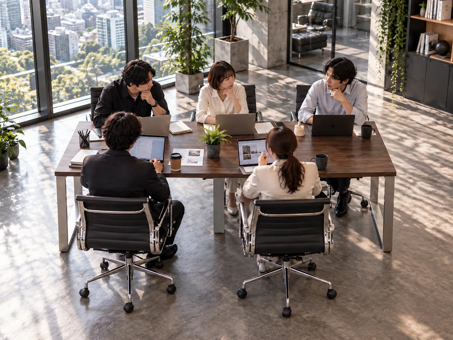 濃い茶色の大きなテーブルで、5人がノートパソコンを開いて打ち合わせをしている会議室